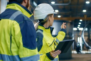 Male and Female Industrial Engineers in Hard Hats and Safety Jackets Discuss New Project while Using Tablet Computer. They Work at the Heavy Industry Manufacturing Factory.