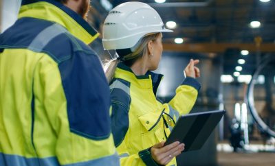 Male and Female Industrial Engineers in Hard Hats and Safety Jackets Discuss New Project while Using Tablet Computer. They Work at the Heavy Industry Manufacturing Factory.