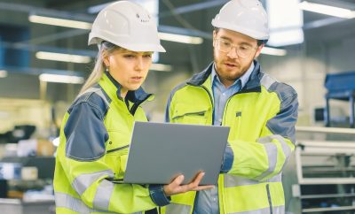 Male Industrial Worker and Female Chief Mechanical Engineer in Walk Through Manufacturing Plant while Discuss Factory's New Project and Using Laptop. Facility Has Working Machinery.