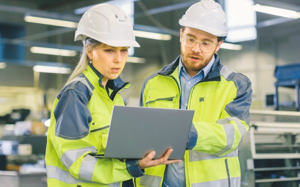 Male Industrial Worker and Female Chief Mechanical Engineer in Walk Through Manufacturing Plant while Discuss Factory's New Project and Using Laptop. Facility Has Working Machinery.